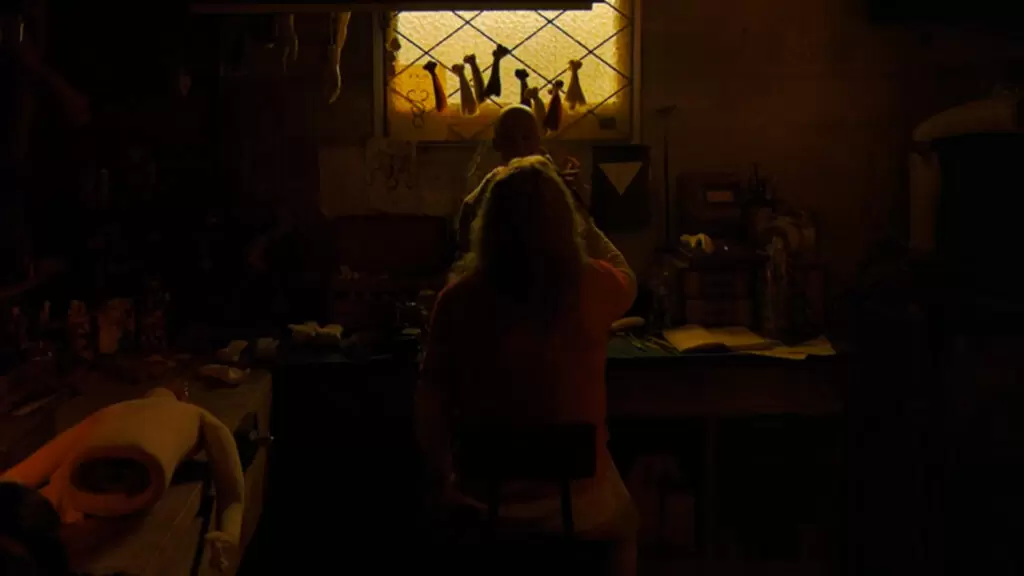 Nicolas Cage como Longlegs. Na cena o ator está de costas e sentado. Ele está usando cabelos longo platinado e confecionando uma boneca de porcelana, em uma sala escura. Apenas com a luz da janela.
